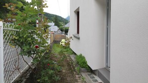A narrow alleyway with a white building on one side and a chain-link fence on the other. There is a small garden area with a few plants and a red flower.