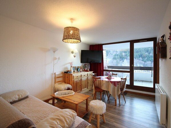 A cozy living room with a sofa, a dining table, and chairs. There is a TV mounted on the wall, and a window with a view of trees.