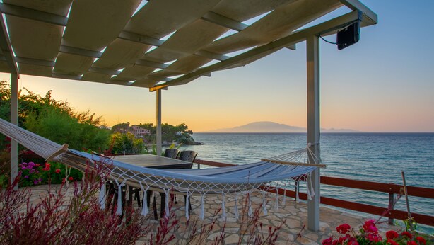 A covered outdoor seating area with a hammock, stone flooring, and a view of the sea. - Baywatch Apartments (Zakynthos)