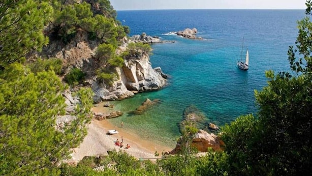 A coastal landscape with a beach, rocky cliffs, and a sailboat in the water. - Leo Street 96, Tossa de Mar-Lesal Villas Rentals Mar Azul (Santa maria de llorell, tossa de mar, gi)