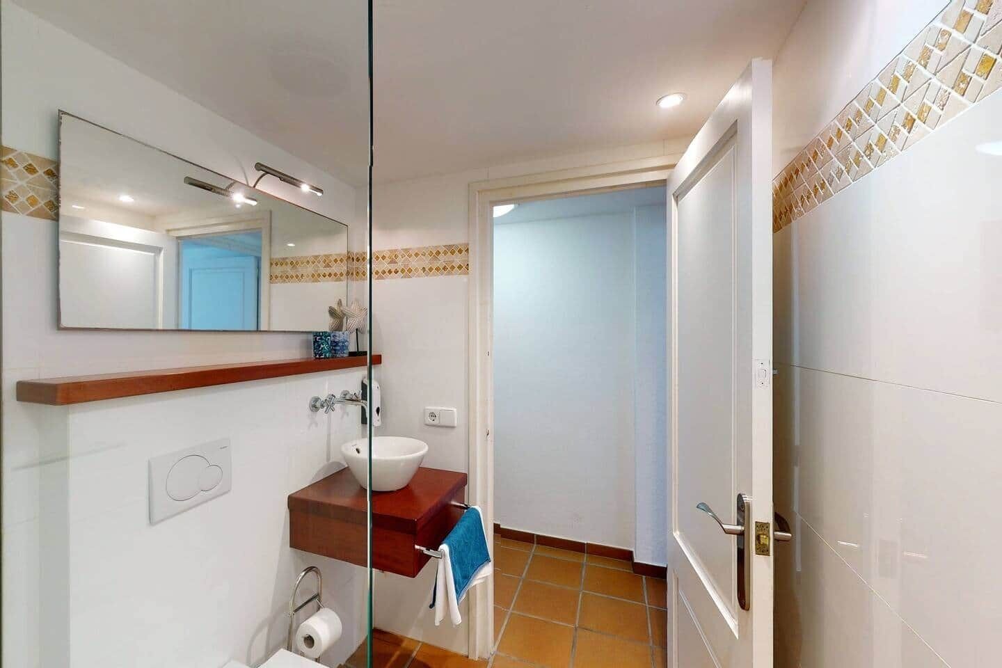 Une salle de bain avec une cloison en verre, un lavabo doté d’un plan de travail en bois, un miroir et une porte donnant sur une autre pièce.