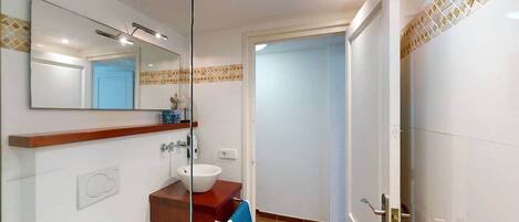 Une salle de bain avec une cloison en verre, un lavabo doté d’un plan de travail en bois, un miroir et une porte donnant sur une autre pièce.