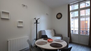 Ein modernes Wohnzimmer mit einem runden Esstisch, grauen Stühlen, einem Garderobenhaken, einer Uhr und Blick auf Gebäude durch große Fenster.