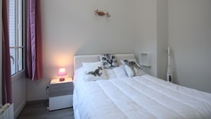 Ein Schlafzimmer mit einem Bett, einem Nachttisch mit Lampe, einem Wandheizgerät und einem Fenster mit Vorhängen.