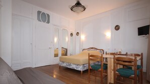 Un dormitorio con una cama, un escritorio y sillas. Hay un armario, un espejo y una lámpara en el techo.