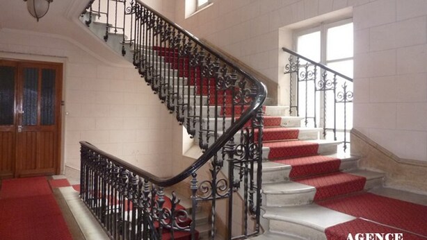 Una imponente escalera con barandillas negras decoradas y peldaños alfombrados en rojo, que conduce a una plataforma con una puerta de madera y un ventanal.
