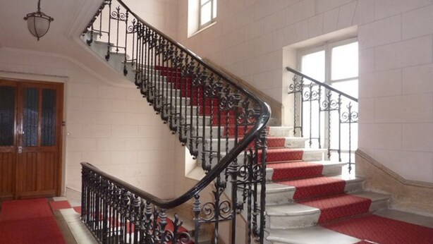 A grand staircase with ornate black railings and red carpeted steps, leading to a landing with a wooden door and a window. - Charming T1 in Mont-Dore facing the Spa, fully equipped, 4 pers. (Mont-Dore)