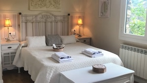Un dormitorio ordenado con una cama grande, dos mesitas de noche con lámparas, una mesita pequeña con una cesta, y una ventana con vistas a la vegetación.