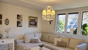Una sala de estar con un sofá color beige, una mesa de centro blanca y una lámpara de araña. Se ven árboles y un edificio a través de las ventanas.