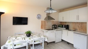 Een kleine keuken met witte kastjes, een eettafel met een plant, een klok en een televisie aan de muur.