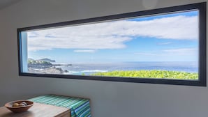 Una habitación con un ventanal amplio que da a la costa, una mesita con un cuenco y un mantel a rayas.