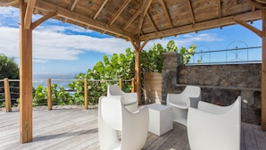 Una terraza de madera con mobiliario exterior blanco, un muro de piedra y vista al mar.
