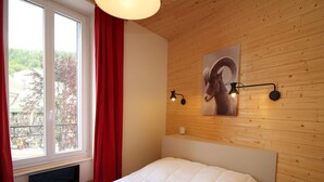 A bedroom with a wooden ceiling, a large window with red curtains, a bed with white bedding, and a mounted picture of a ram's head.