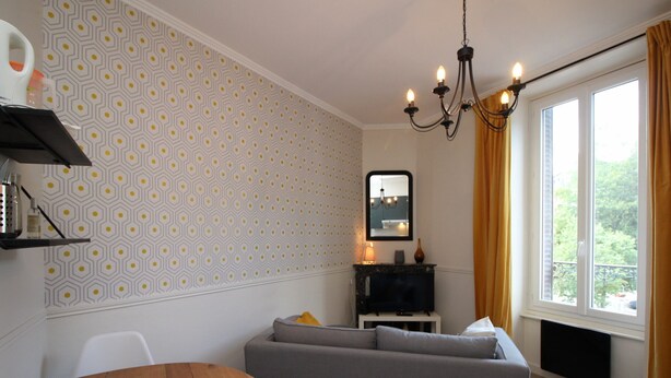 A modern living room with a grey sofa, a wooden table, a patterned wallpaper, a chandelier, and a large window with curtains.