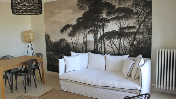 Un salon avec un canapé blanc, une table à manger en bois avec des chaises, un luminaire suspendu noir en fibres tressées, une lampe murale et un grand tableau représentant un arbre en noir et blanc.