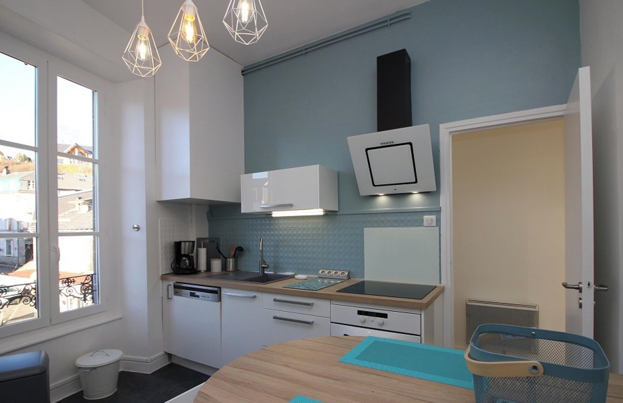 A modern kitchen with white cabinets, a wooden countertop, and a blue backsplash. There is a window providing natural light, and a door leading to another room.