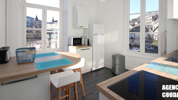 Eine moderne Küche mit weißen Schränken, einer Mikrowelle, einem Kühlschrank und einem Spülbecken. Zwei Fenster bieten Blick auf die Stadt.