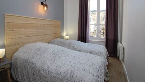 Ein Schlafzimmer mit zwei Betten, einem Holzkopfende, einem Nachttisch mit Lampe, einem Fenster mit Vorhängen und Blick ins Freie.