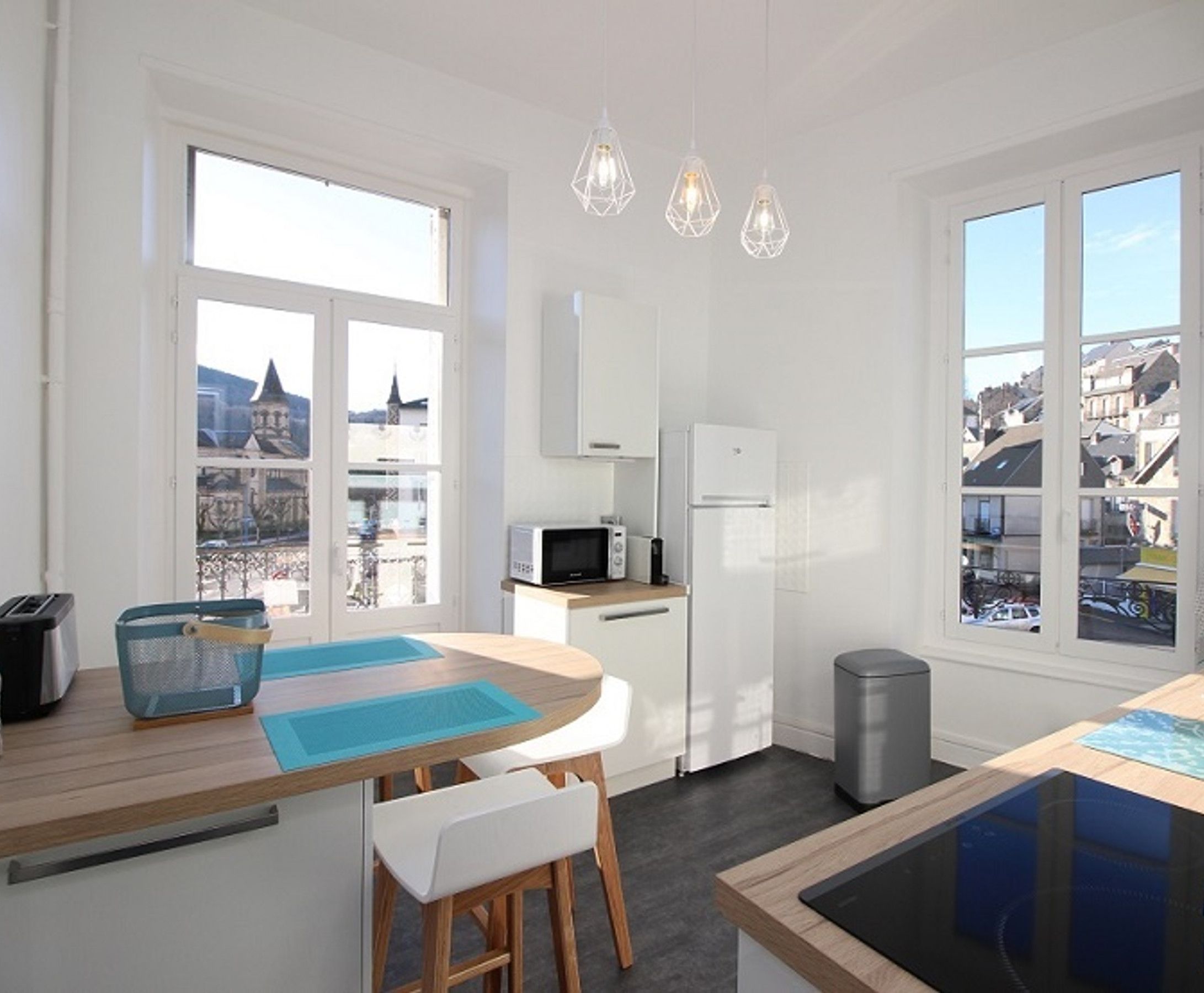 A modern kitchen with white cabinets, a microwave, a refrigerator, and a sink. There are two windows providing views of the city.