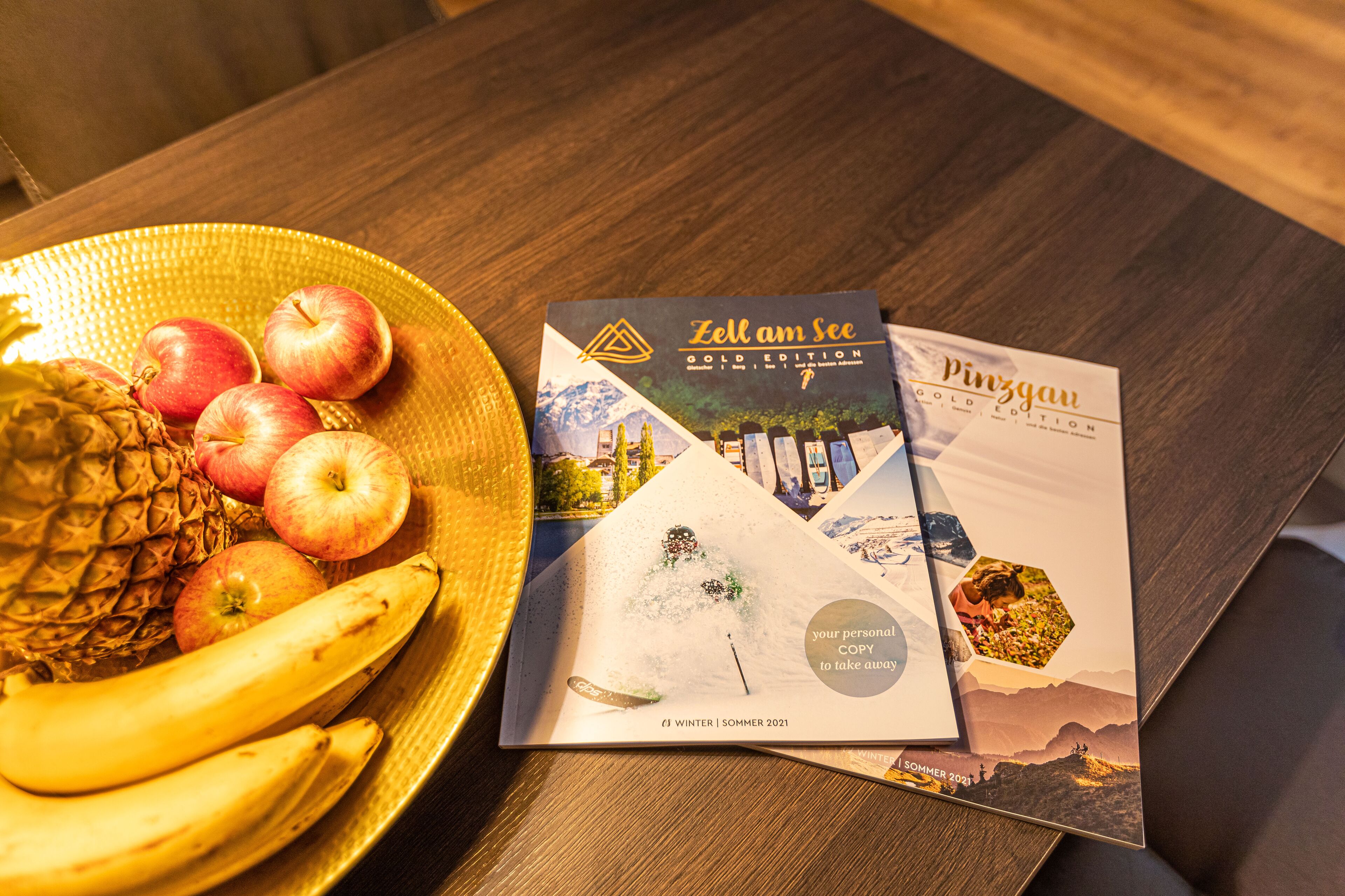 A table with a fruit bowl containing bananas, apples, and a pineapple, and two brochures titled "Zell am See Gold Edition" and "Pinzgau".