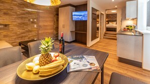 A modern hotel room with a wooden dining table, a fruit bowl, a bottle of wine, and a magazine. - Tevini Boutique Suites by we rent (Zell am See)