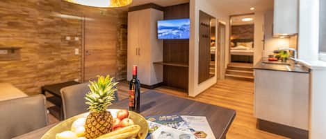 A modern hotel room with a wooden dining table, a fruit bowl, a bottle of wine, and a magazine.
