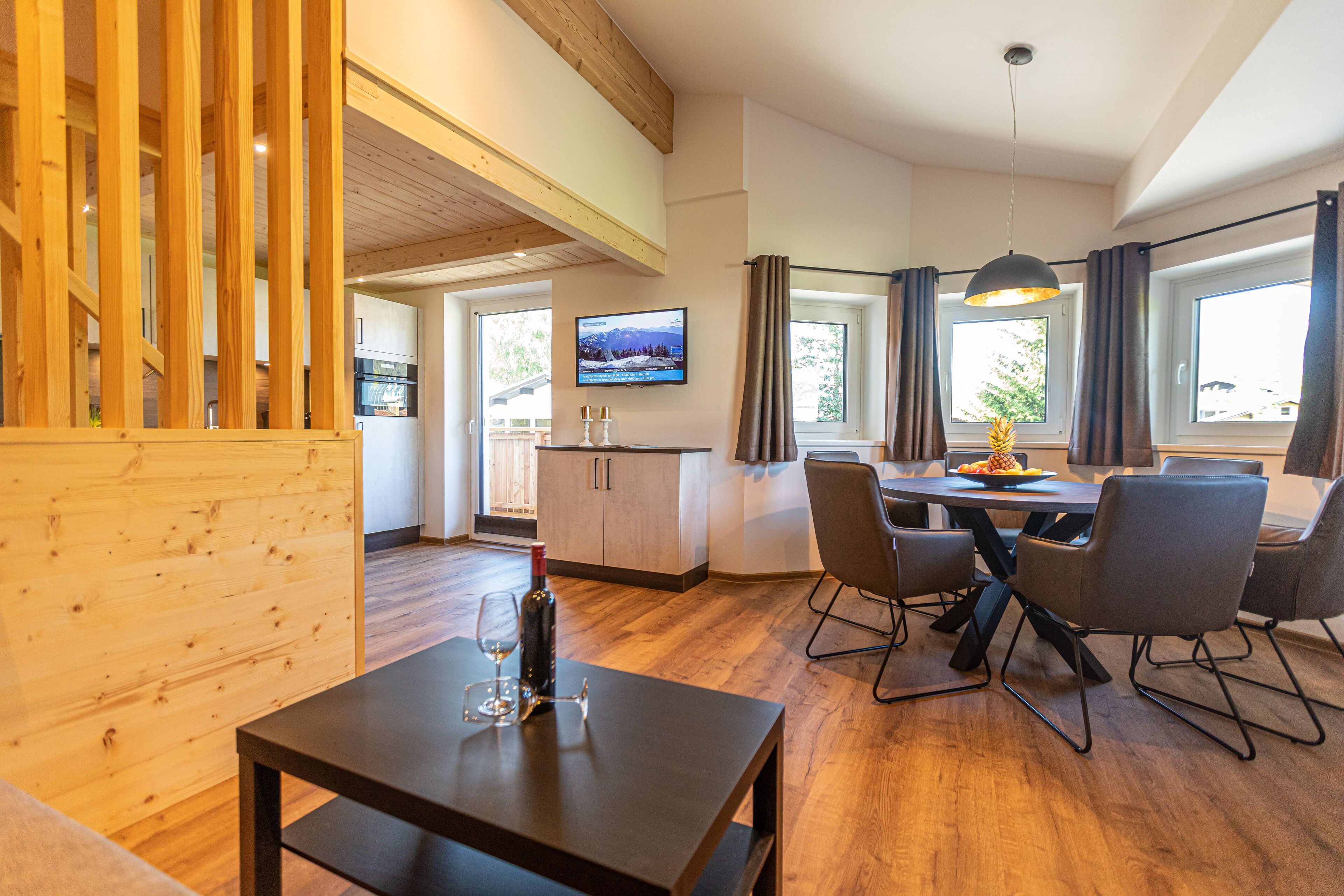 Een moderne keuken met houten kastjes, een eethoek met een tafel en stoelen, en een televisie aan de muur.