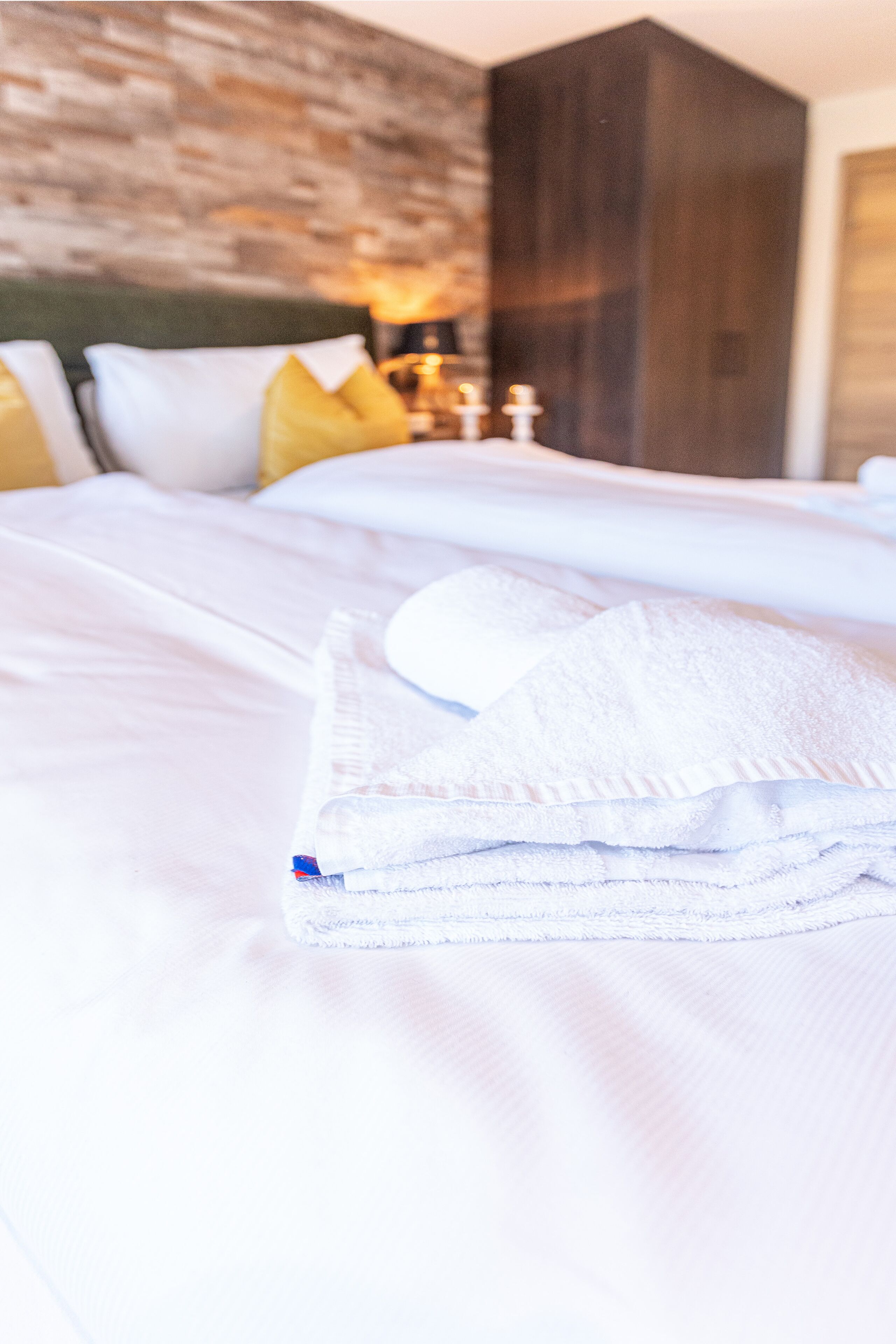 A neatly made bed with white linens and a folded towel, a wooden headboard, and a wall with a textured pattern.