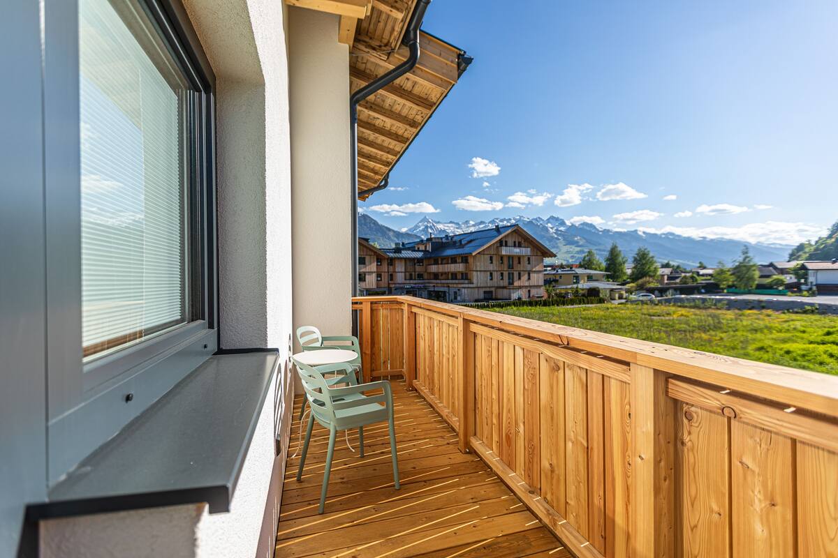 Un balcon avec une balustrade en bois, une fenêtre avec des stores et deux chaises.