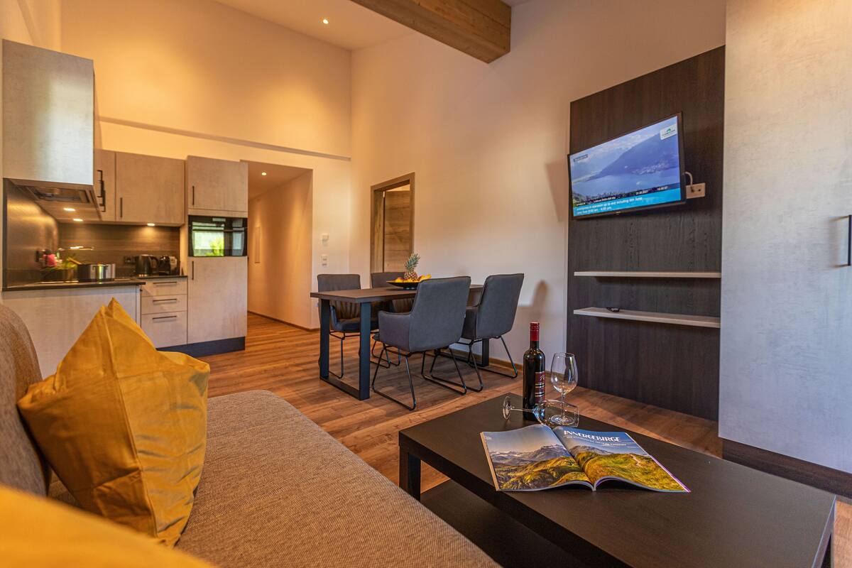 Un salon moderne comprenant un canapé, une table basse avec un magazine, un téléviseur fixé au mur et un coin repas avec des chaises et une table.