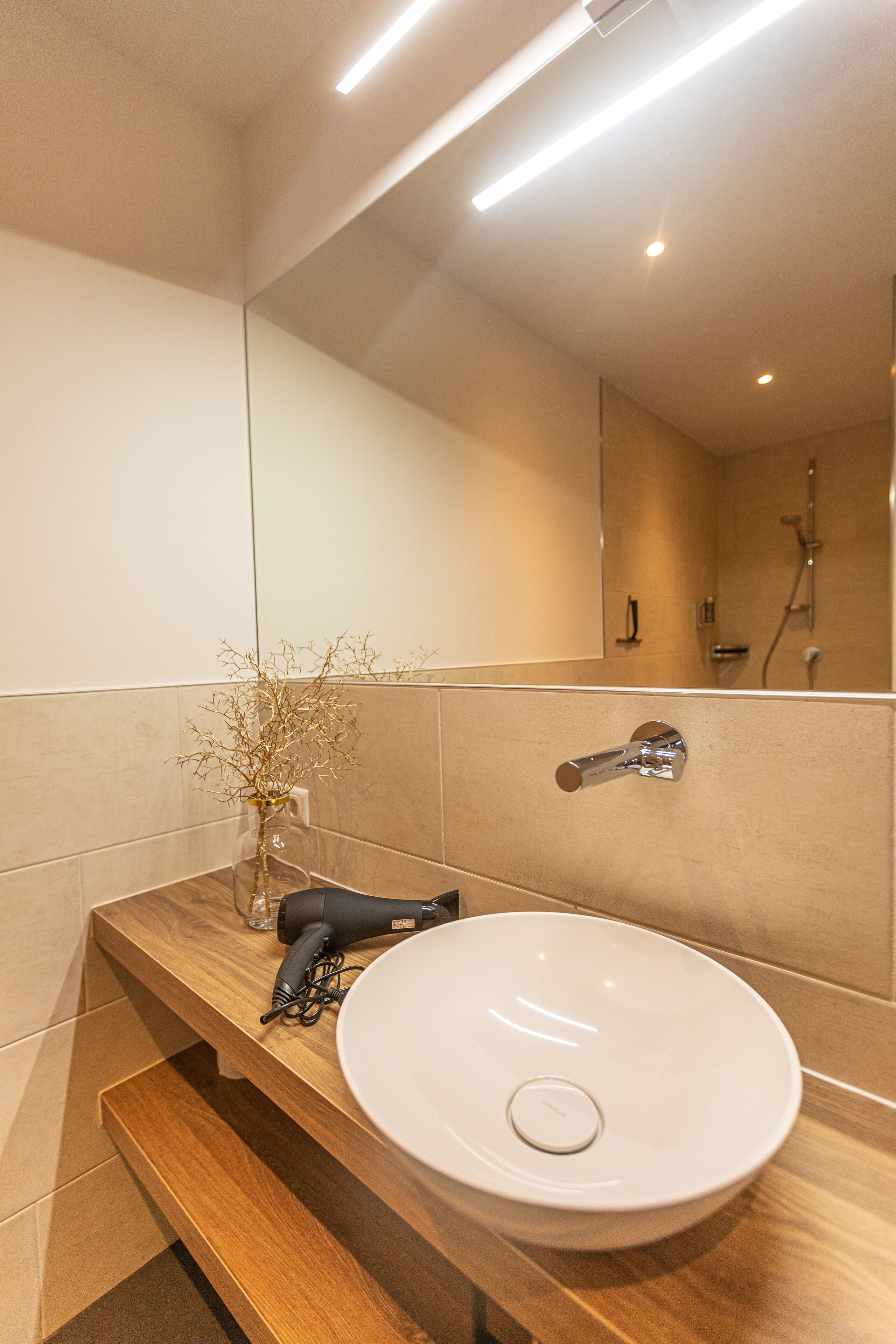 A modern bathroom with a wooden vanity, a white sink, a hairdryer, and a vase with dried branches.