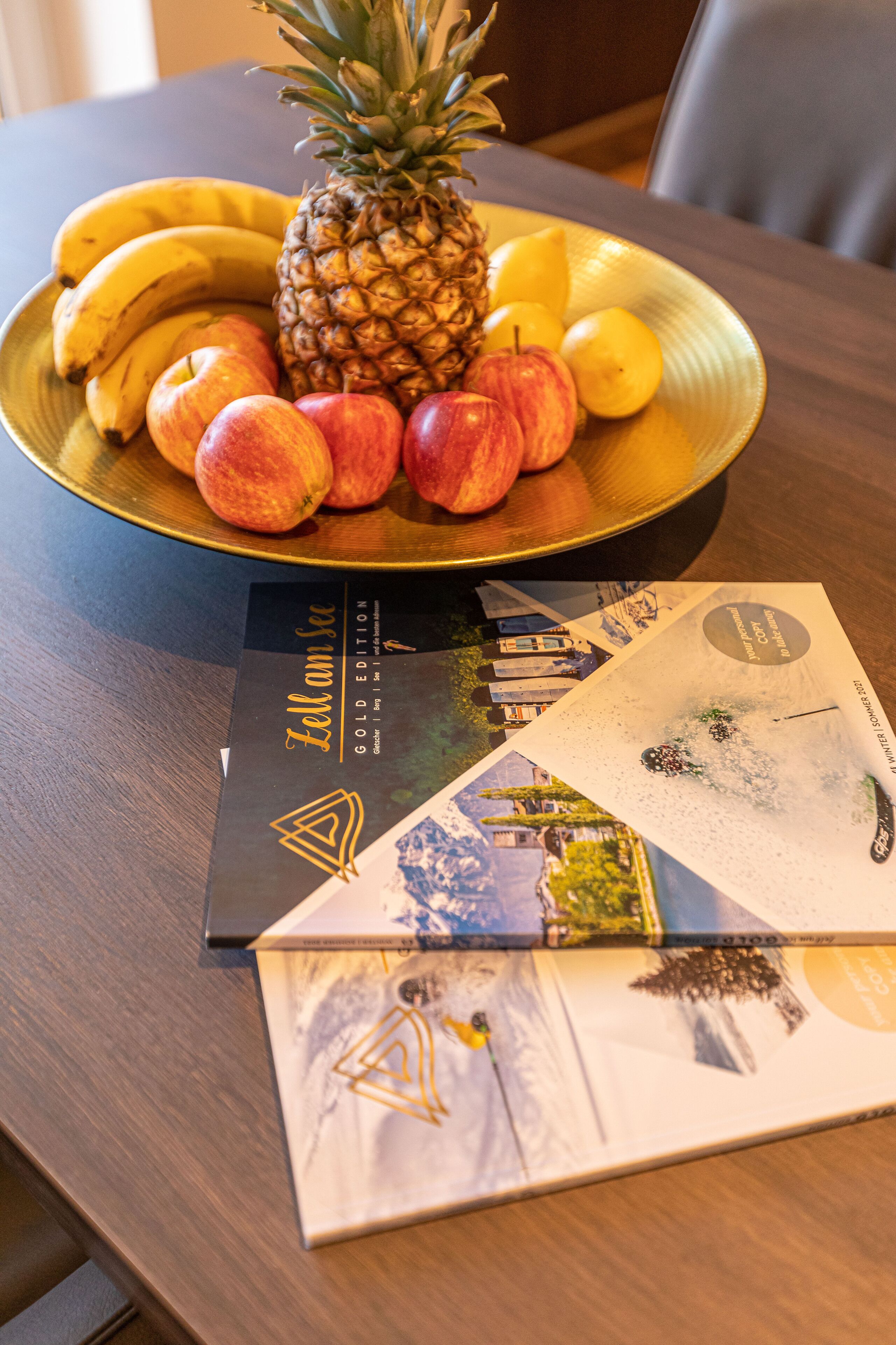 Una mesa de comedor con un frutero que contiene plátanos, manzanas y una piña, y una revista titulada 'Zell am See'.