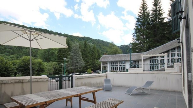 Une terrasse extérieure avec des tables et des bancs en bois, un grand parasol et une vue sur un bâtiment à la façade blanche, avec des arbres verdoyants en arrière-plan.