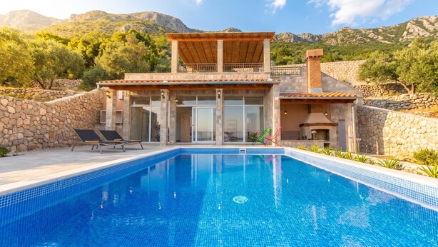 A poolside villa with a stone wall, a covered patio, and a mountain backdrop.