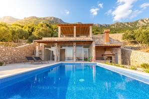 Una villa junto a la piscina con muro de piedra, porche cubierto y montañas al fondo.