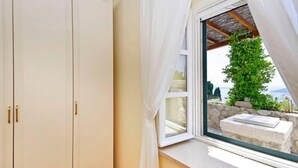 A room with a large window, a wardrobe, and a view of a stone wall and greenery outside.