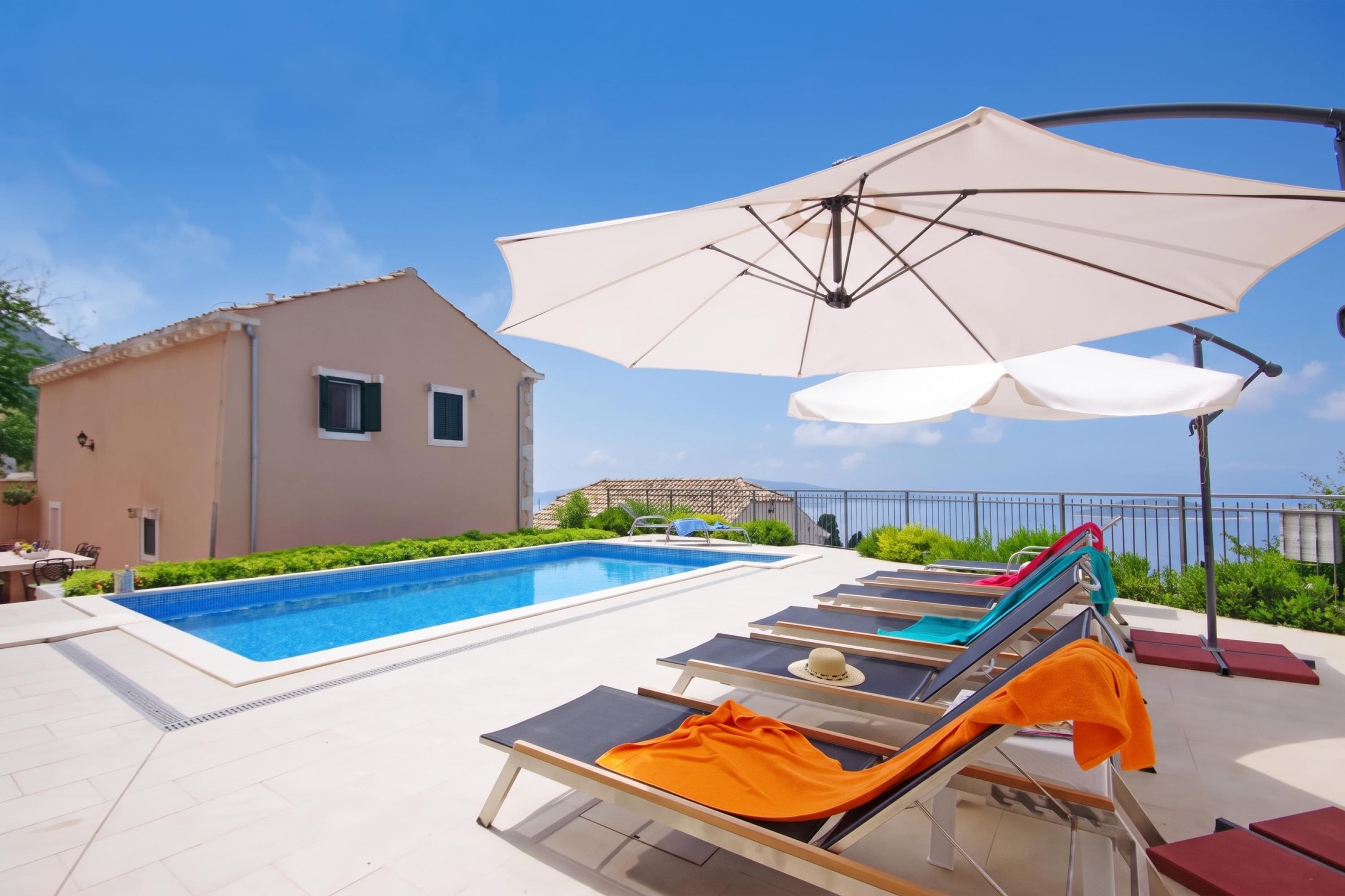 A swimming pool with lounge chairs and umbrellas, a building with a tiled roof, and a view of the sea.