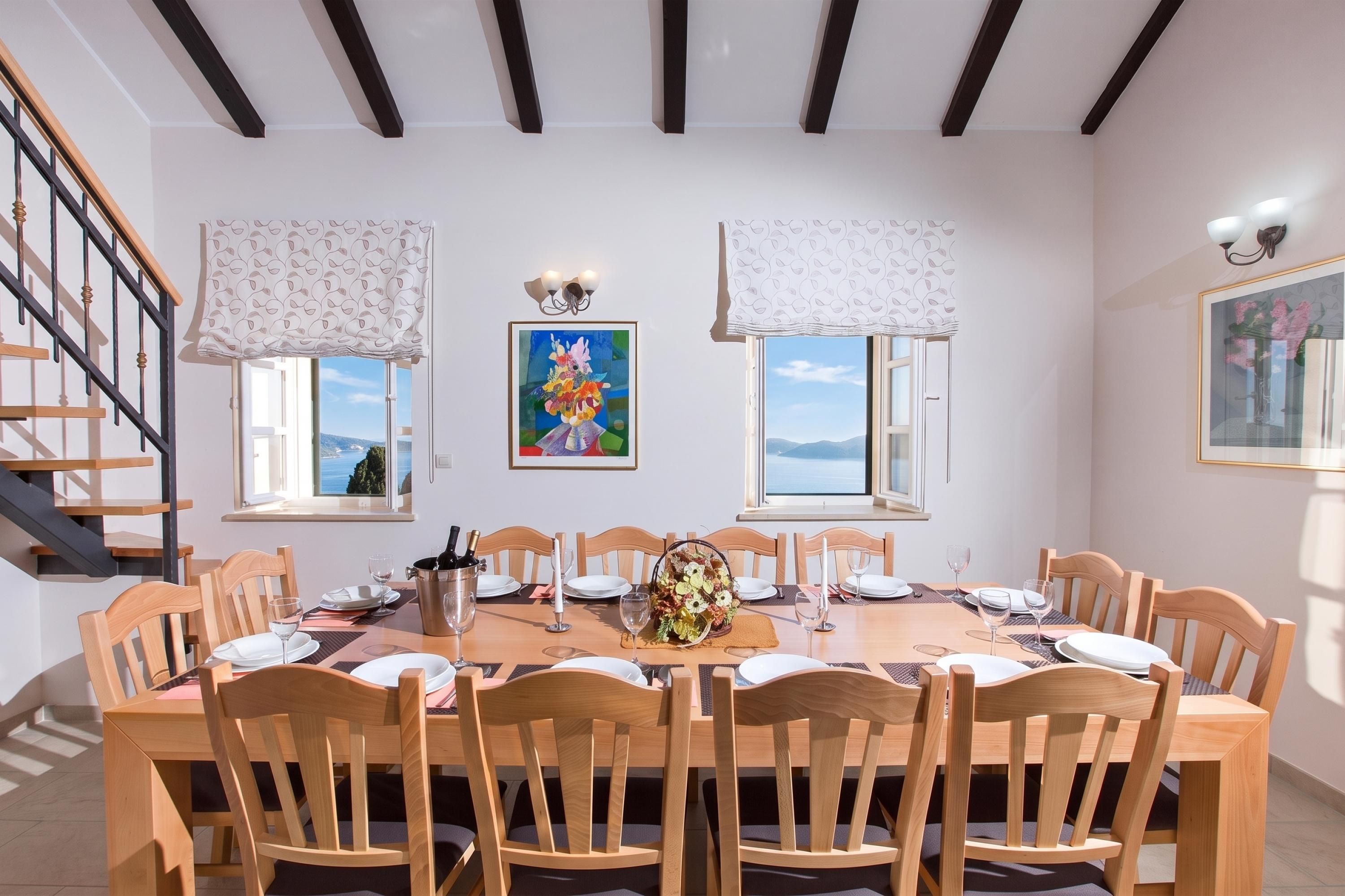 A dining room with a wooden table set for six, flanked by two windows with white curtains, a painting above the windows, and a staircase on the left.