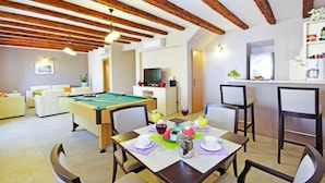 A room with a pool table, dining table with chairs, a kitchen area, and a living space with a sofa.
