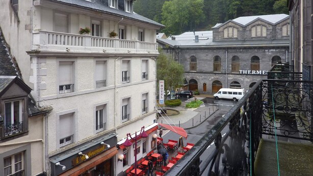Un hotel con balcón, un restaurante con terraza y un edificio con la leyenda 'THERMES' al fondo.
