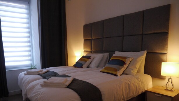 A neatly made bed with pillows and a blanket, a bedside table with a lamp, and a window with blinds. - Baxter Park City Base (Dundee)