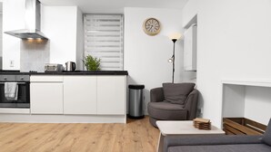 A modern kitchen with white cabinets, a stainless steel range hood, and a black countertop. There is a trash can, a clock on the wall, and a grey sofa in the living area. - Baxter Park City Base (Dundee)