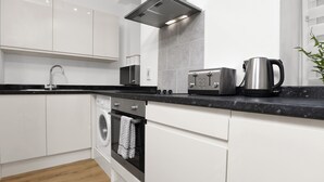 A modern kitchen with white cabinets, a stainless steel range hood, a built-in oven, a washing machine, and a coffee maker. - Baxter Park City Base (Dundee)