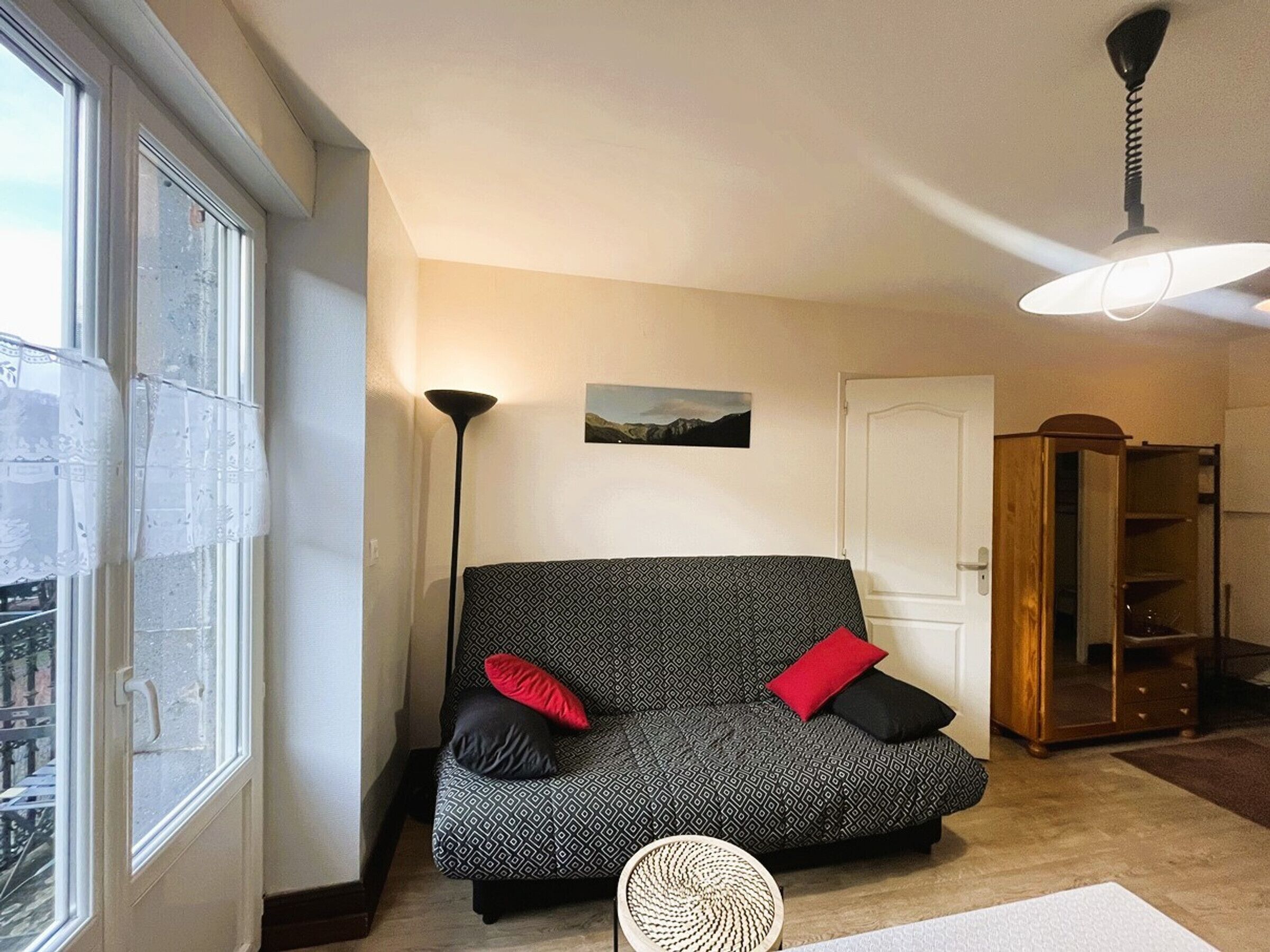 A living room with a sofa, a standing lamp, a ceiling fan, a door, and a window with a view of buildings.