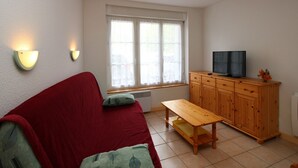 A room with a red sofa, a wooden cabinet with a TV, and two wall-mounted lights. - Mont-Dore: 2-room Apartment near Thermal Baths, Sleeps 4 (Mont-Dore)