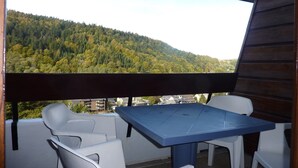 A balcony with a blue table and white chairs, overlooking a forested hillside. - Mont-Dore - Studio with balcony, parking, wifi included (Mont-Dore)
