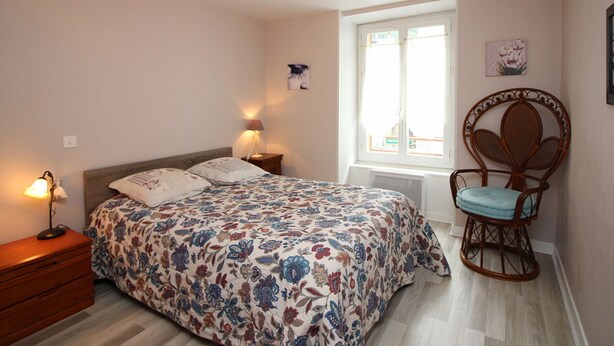 A bedroom with a bed, a chair, a nightstand, a lamp, a window, and a wall with a picture. - Central T2 near spa with wifi (Mont-Dore)