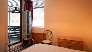 A bedroom with a bed, a chair, a wooden dresser, a window with blinds, and a clothes rack. - Mont-Dore: 1BR with Balcony, Covered Parking, 4 pax (Mont-Dore)