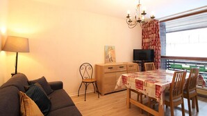 A cozy living room with a sofa, a dining table with chairs, a TV, and a chandelier. - Mont-Dore: 1BR with Balcony, Covered Parking, 4 pax (Mont-Dore)
