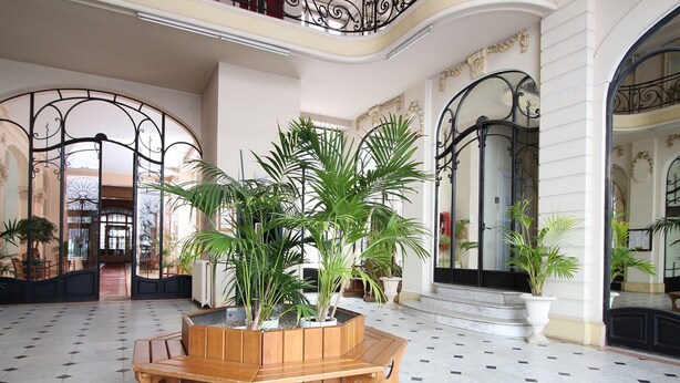 A spacious lobby with a central wooden bench, potted plants, and a tiled floor. - Charming city center studio with wifi (Mont-Dore)